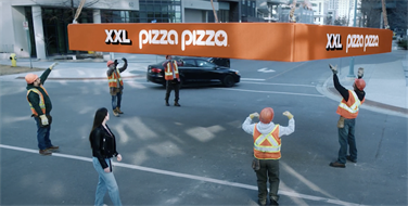 A group of construction workers reaching up to grab a giant pizza box being lowered into an intersection 